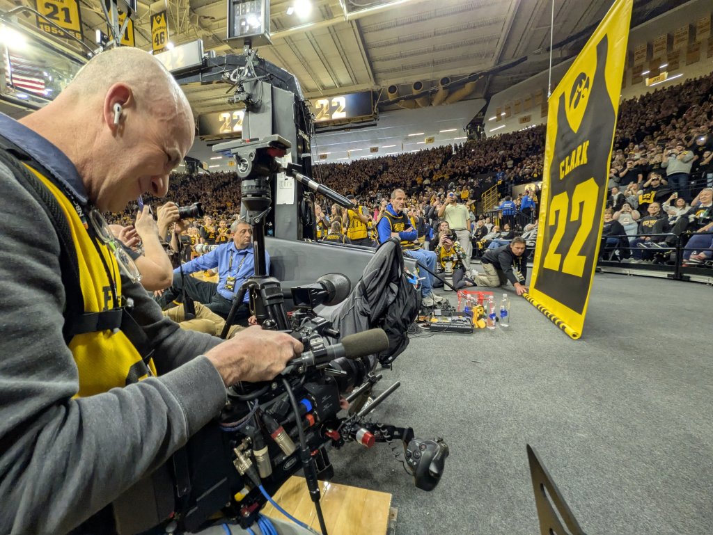 Photograph taken from the floor of Carver Hawkeye Arena as a banner of Caitlin Clark's #22 jersey is lifted towards the ceiling of the arena, retiring her number. A photographer is on the left side of the frame recording the event.